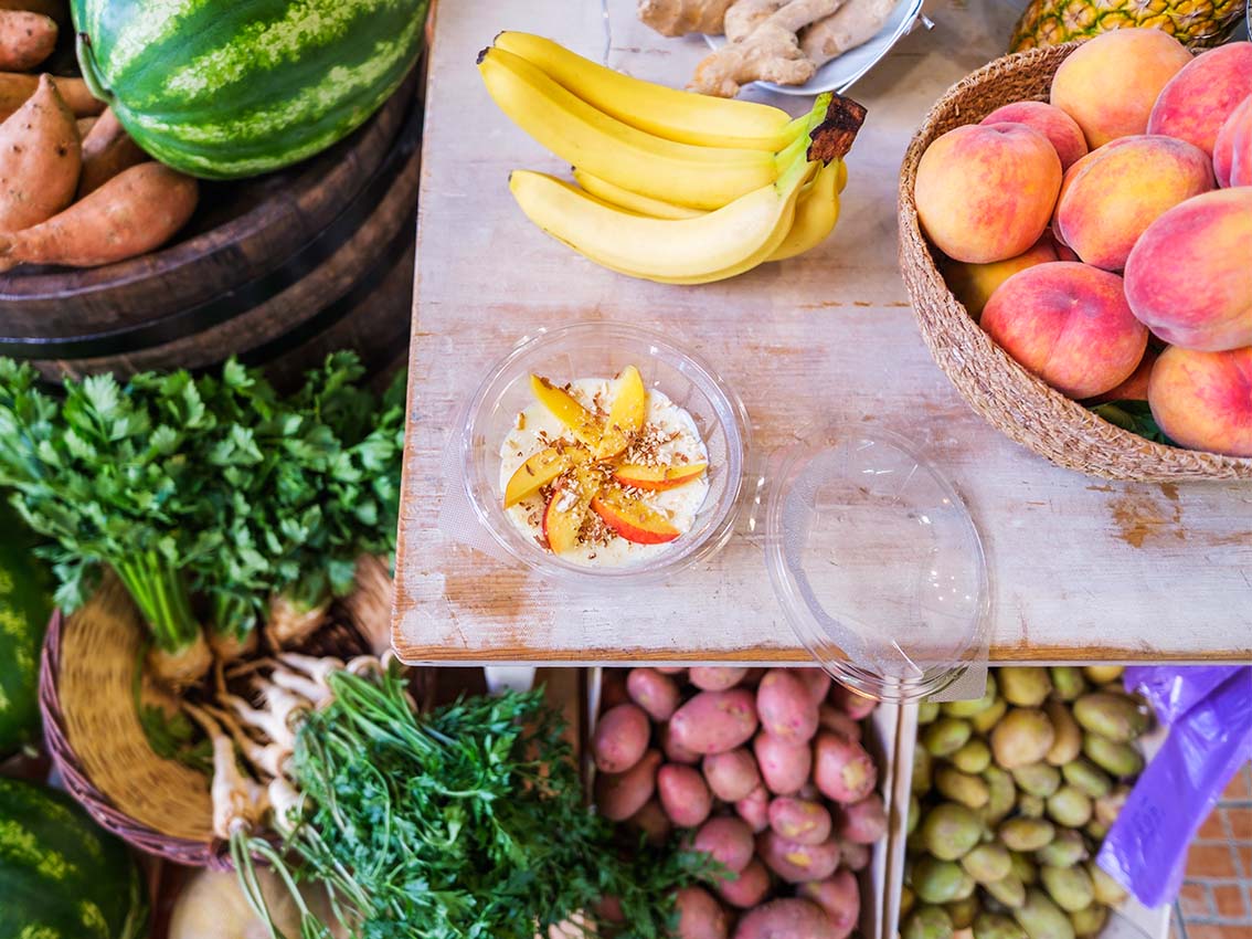 Mesa com frutas como banana, pêssego e mamão, ao fundo melancias e batatas